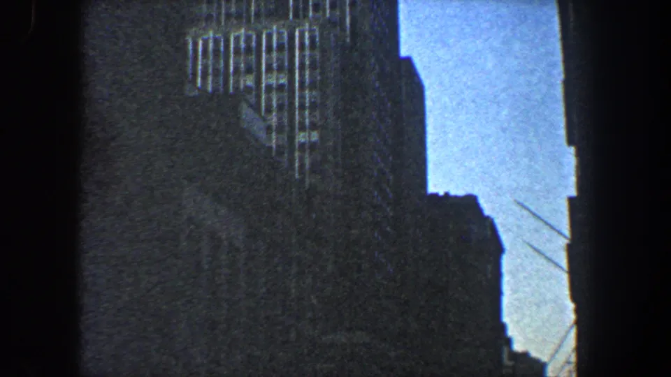 1960 New York City 8mm frame showing tall office-building facades and a narrow blue sky gap from below.