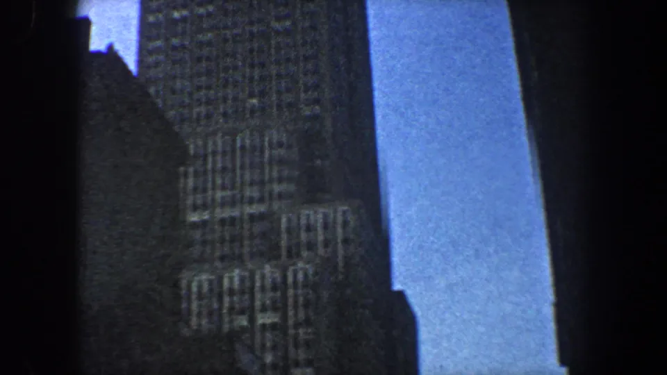 1960 New York City 8mm frame of a gridded high-rise facade beside dark building silhouettes and open blue sky.
