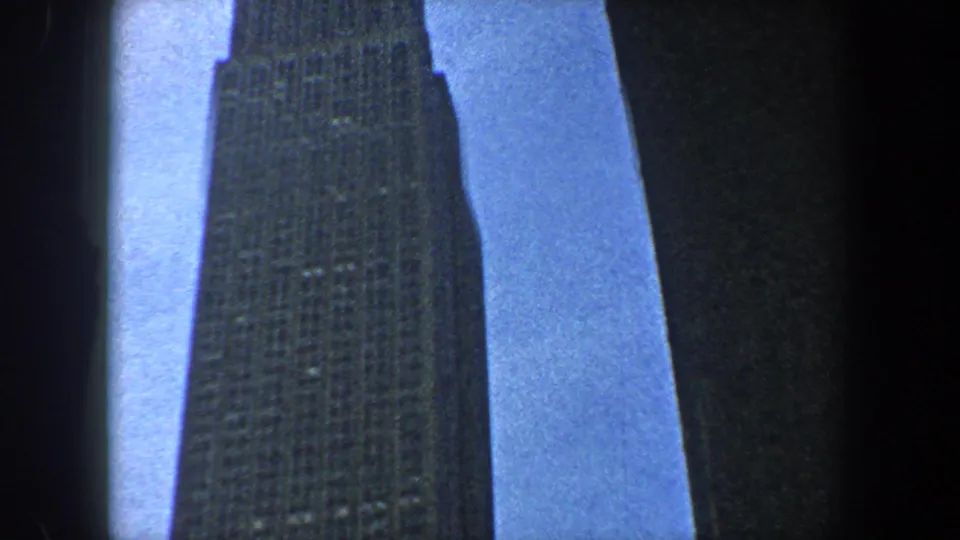 1960 8mm footage shows a tall New York skyscraper viewed from below, with blue sky and dark neighboring building edges.