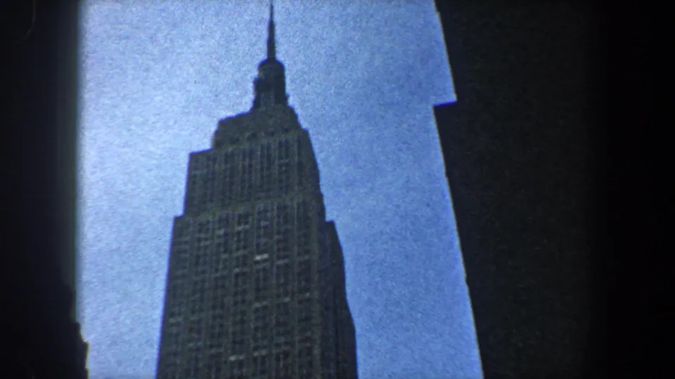 The Empire State Building's upper tower and spire appear in 1960 New York City footage, framed by darker neighboring skyscrapers.