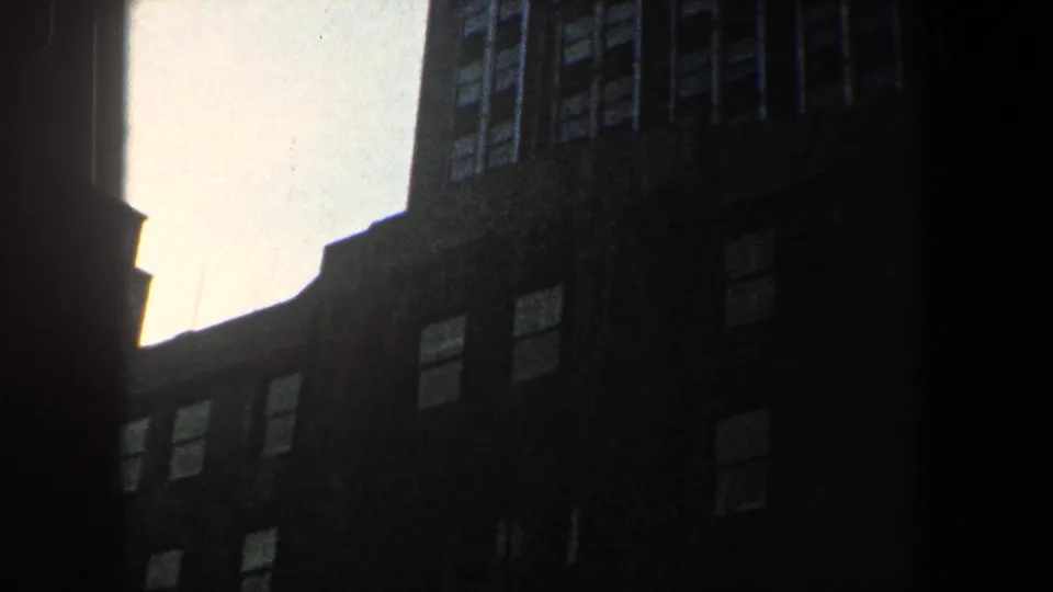 1960 New York City home-movie frame of a shadowed high-rise wall with window rows beneath a pale sky.