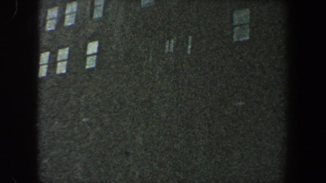 Dark urban building facade with rows of bright windows and heavy film grain.