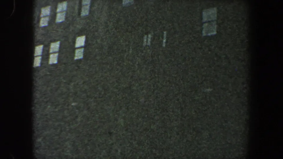 1960 New York City 8mm frame showing a dark building exterior with scattered bright window reflections and visible gate-edge shadow.