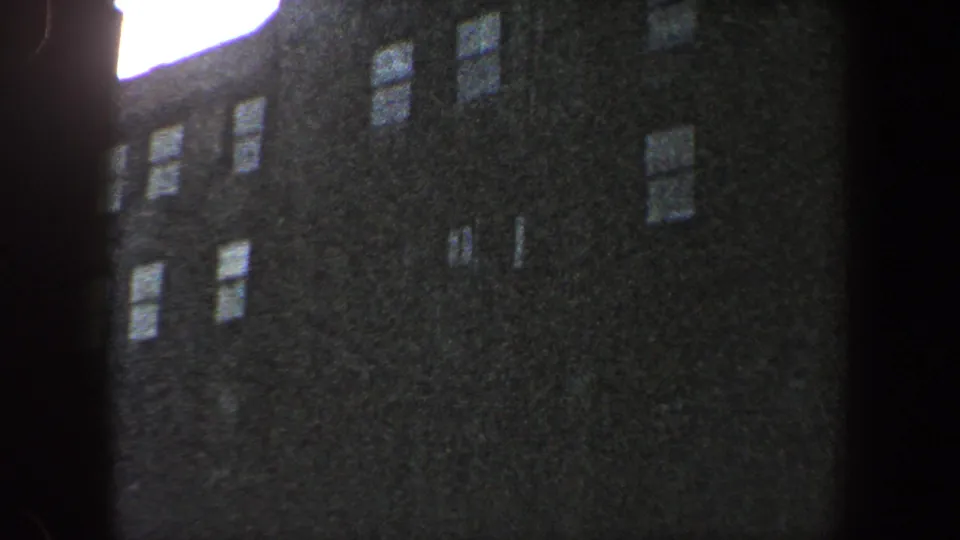 1960 New York City 8mm view of a tall shadowed building, bright sky flare at the corner, and several small window openings.