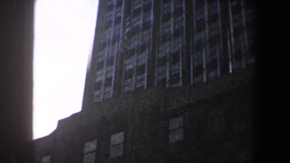 1960 New York City 8mm frame looking upward between dark building exteriors, with bright sky and vertical window rows.