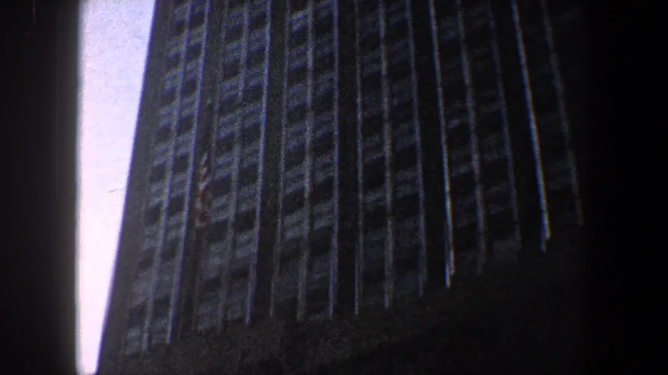 1960 New York City home-movie frame showing a low-angle view up a tall gridded skyscraper facade against pale blue sky.