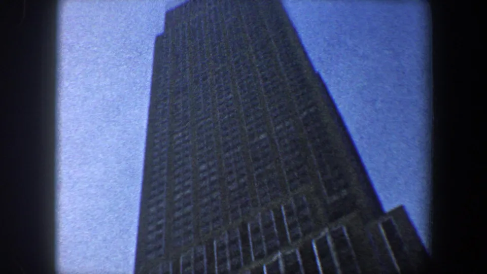 1960 New York City home-movie frame looking up along a tall skyscraper shaft, with gridded windows and blue sky at the edge.
