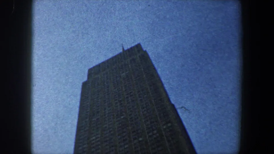 Low-angle 1960 New York City frame showing the upper portion of an unidentified skyscraper against a grainy blue sky.