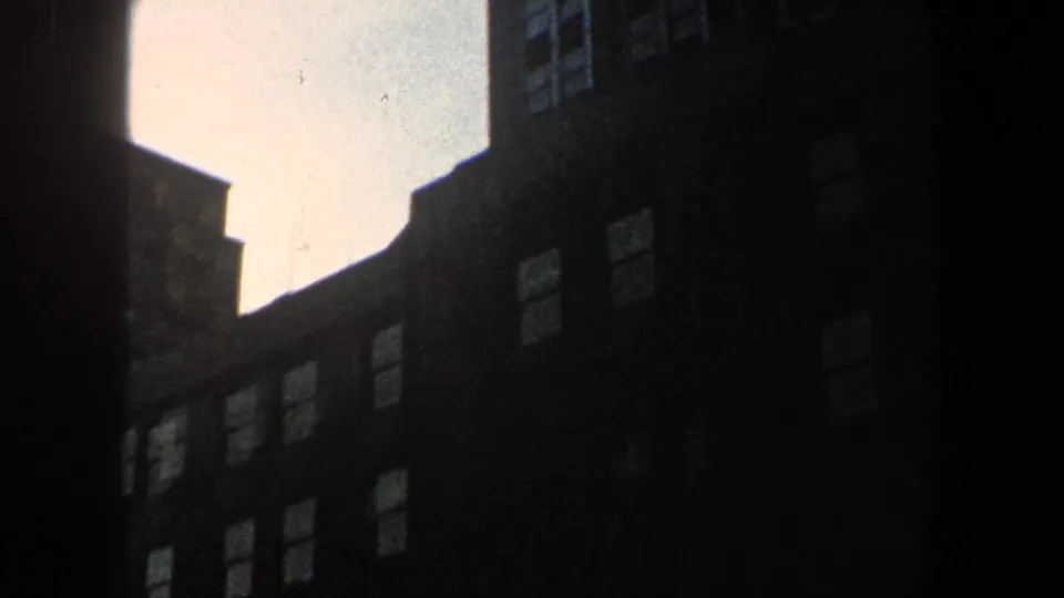 1960 New York City 8mm frame looking upward between shadowed building facades toward a pale sky.