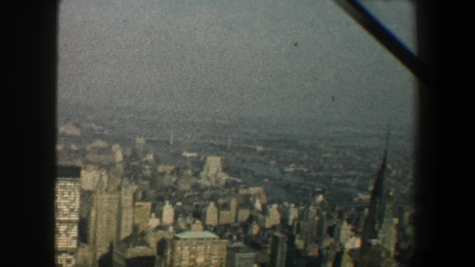 1960 Manhattan cityscape from above, showing dense buildings, a distant waterway, haze, and a spire-like tower near the lower right.
