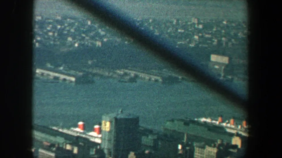 1960 New York City harbor view with piers, ships, waterfront buildings, and a distant shoreline seen through diagonal tower framing.