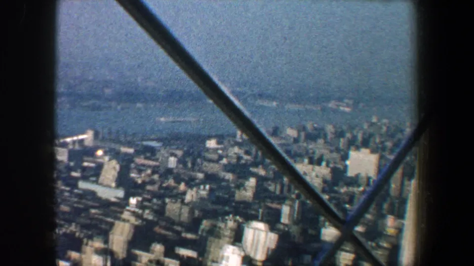 1960 New York City aerial view with high-rises, harbor water, haze, and dark diagonal beams in the foreground.