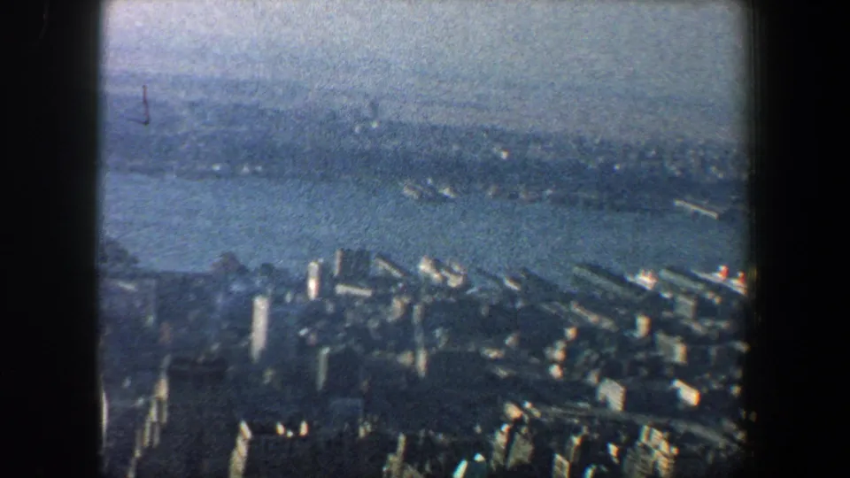 1960 New York City 8mm frame showing a hazy aerial sweep over riverfront buildings, piers, and water toward a distant shore.