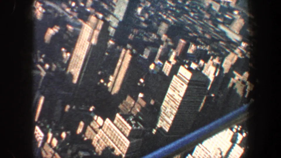 1960 New York City aerial frame with tilted dense blocks, bright high-rise facades, and a blue structural or frame edge crossing below.