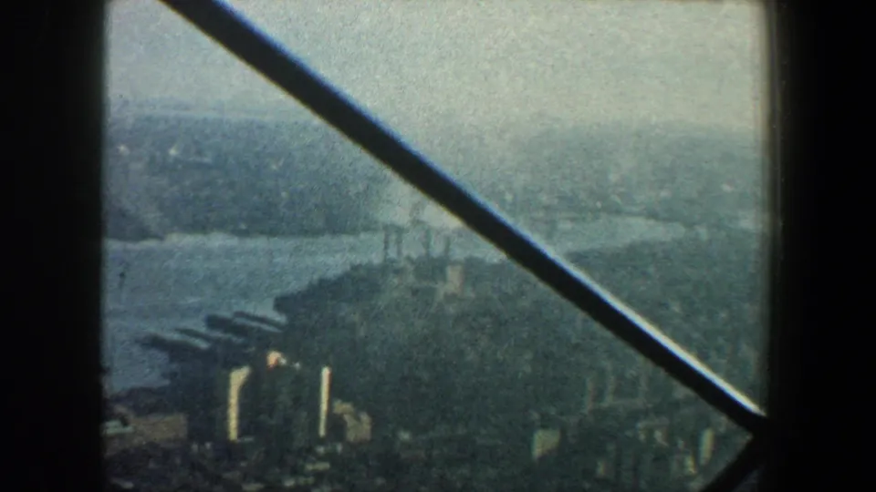 New York City waterfront and distant urban blocks seen from above, with a dark diagonal support beam crossing the frame.