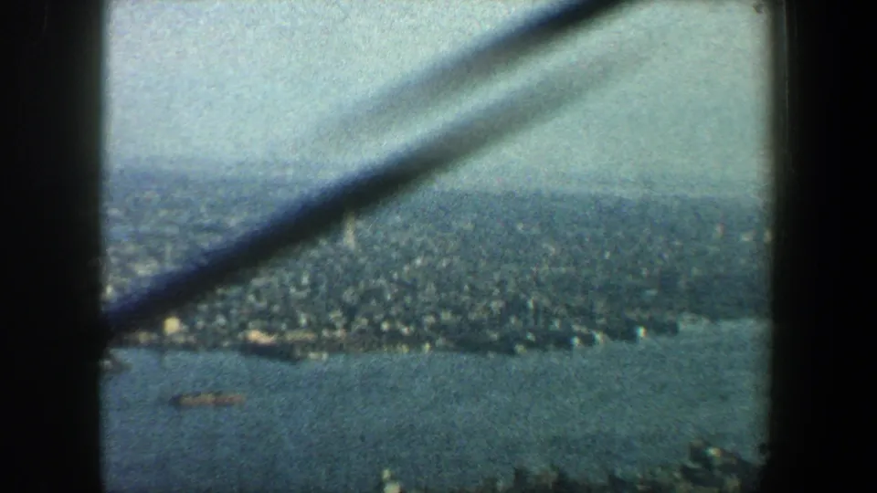 1960 New York City aerial view through aircraft window framing, with harbor water, shoreline blocks, and haze in the distance.