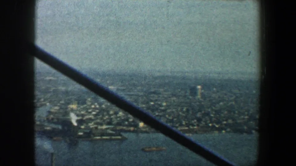 Aerial New York City waterfront scene from 1960, with piers, urban blocks, water, and aircraft structure crossing the frame.