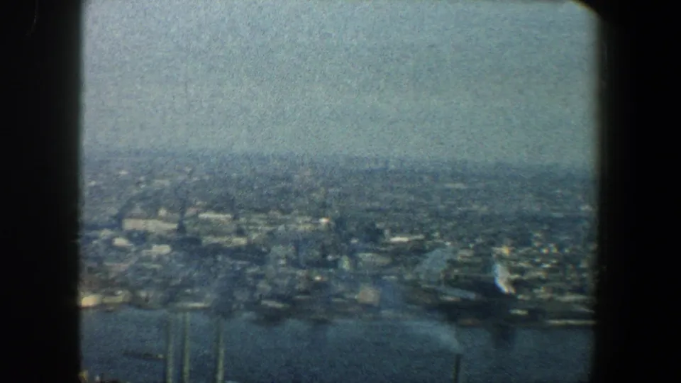 Soft 8mm aerial view over New York City in 1960, showing waterways, waterfront structures, and a hazy spread of buildings.