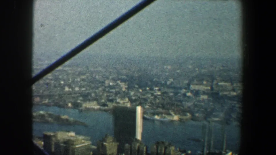 New York City aerial home-movie frame from 1960 with water, clustered buildings, a tall light-colored tower, and aircraft window posts.