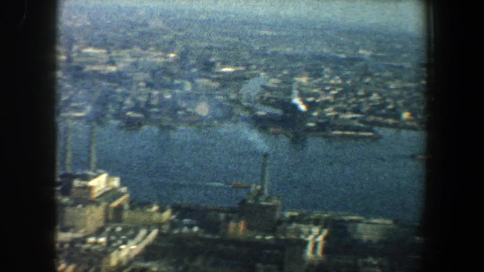 1960 New York City home-movie frame looking across river water, piers, tall buildings, and a broad urban shoreline.