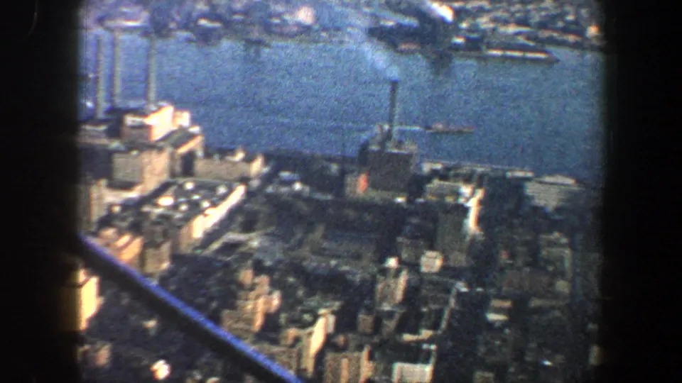 Elevated 1960 New York City waterfront view with buildings, piers, river, and crossing observation-deck beams in soft 8mm footage.