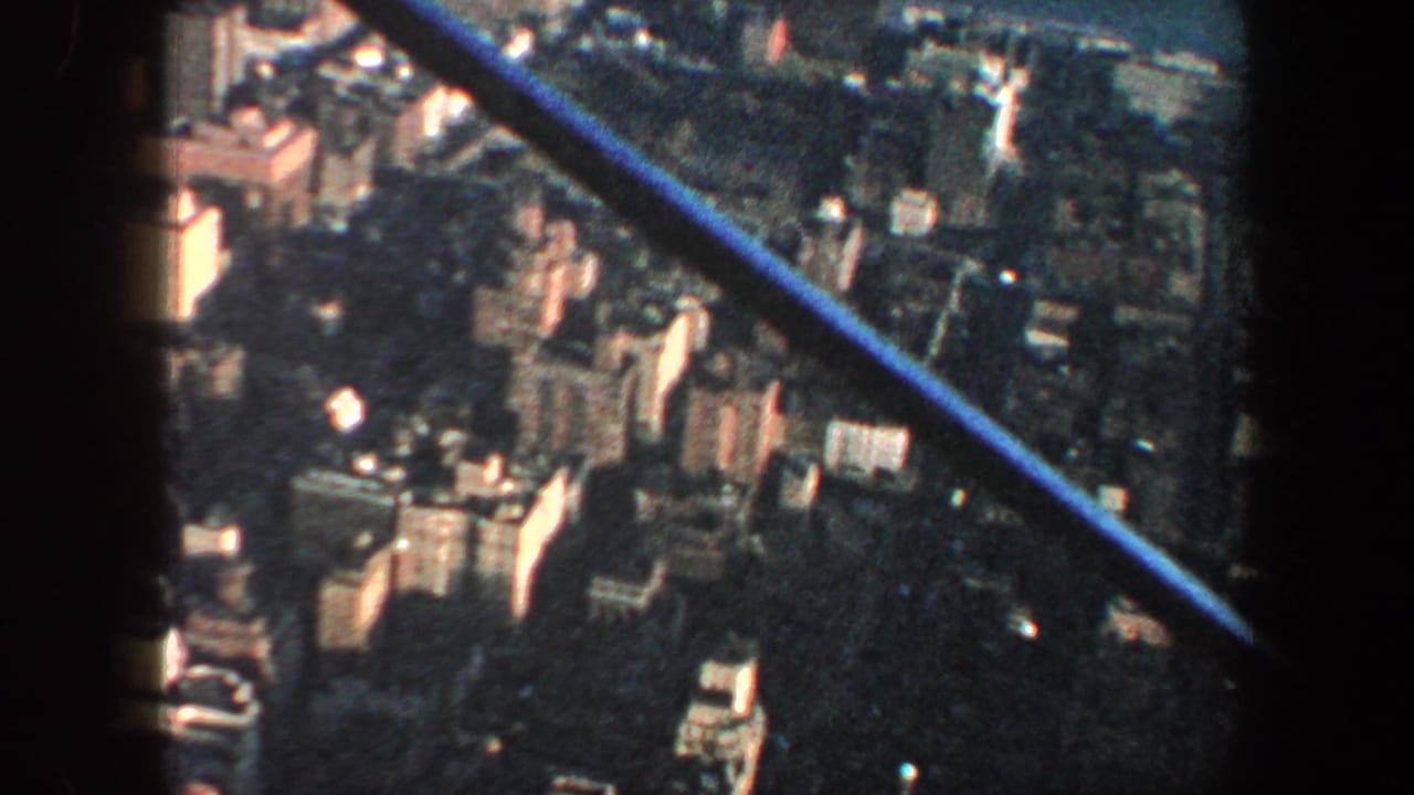 High aerial view of dense urban blocks with a blue diagonal aircraft strut crossing the frame.