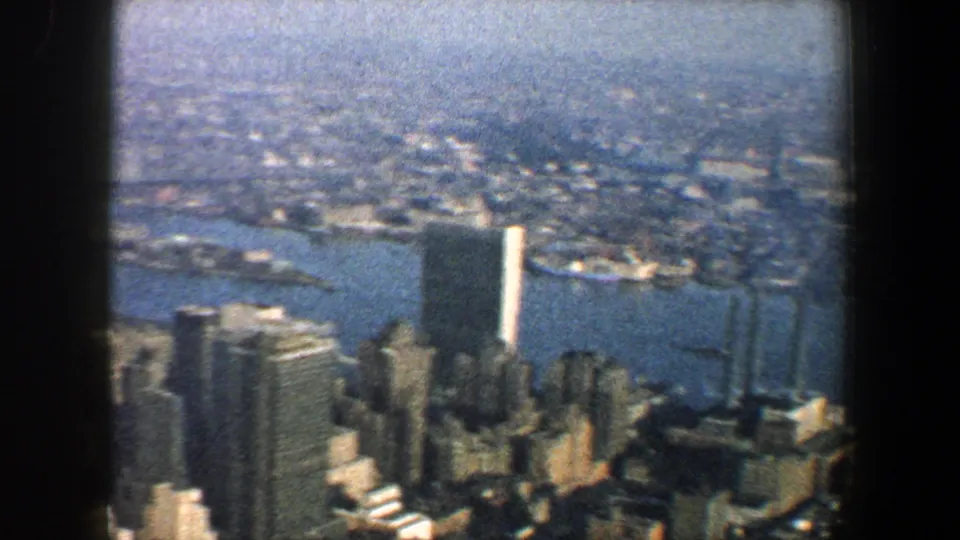 1960 New York City skyline viewed from above through dark window framing, with riverfront piers and tall buildings visible.