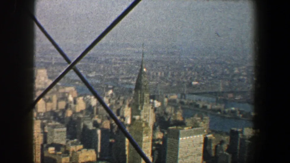 New York City skyline from above with a pointed skyscraper spire, river in the distance, and diagonal observation-window bracing.