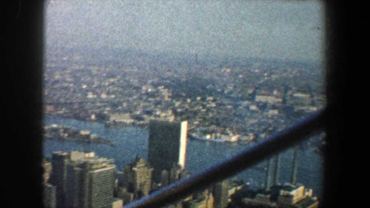 1960 8mm view over New York City buildings and waterfront from a high structure, crossed by dark vertical and diagonal beams.
