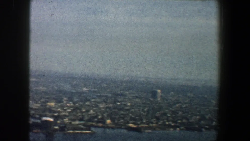 Soft 8mm frame of New York City from above, showing waterfront blocks, distant buildings, and a pale hazy sky.