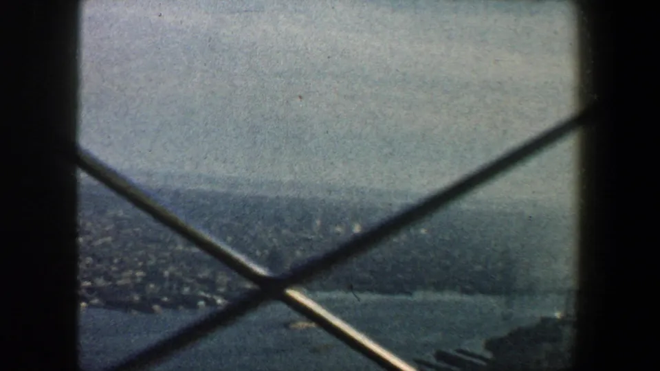 High 1960 New York City view through crossing tower beams, with river, shoreline, and distant urban forms in haze.