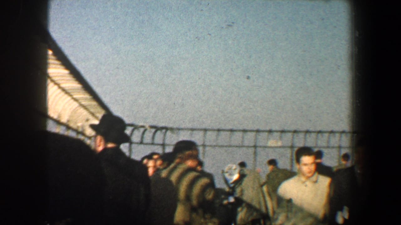 Crowd in coats standing behind a barbed wire-topped fence under a roof edge.