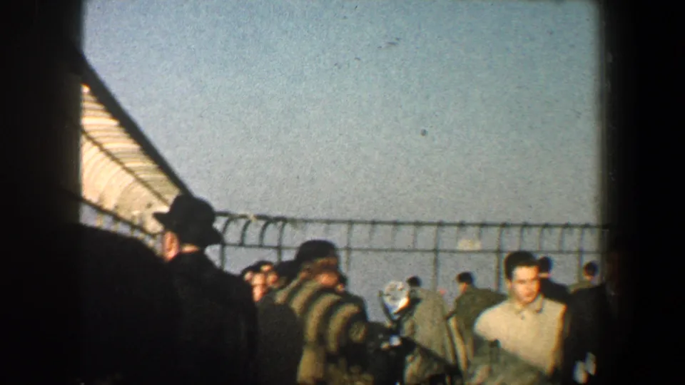 1960 New York City home-movie frame showing adults in coats and hats near a barbed wire-topped fence with sky and water beyond.