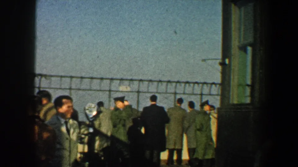 1960 New York City home-movie frame showing a small crowd facing a fenced overlook, with coats, hats, and a distant pale background.