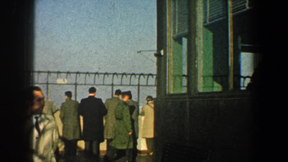 1960 New York City home-movie frame showing adults near a green structure and barbed wire-topped fence, with an open view beyond.