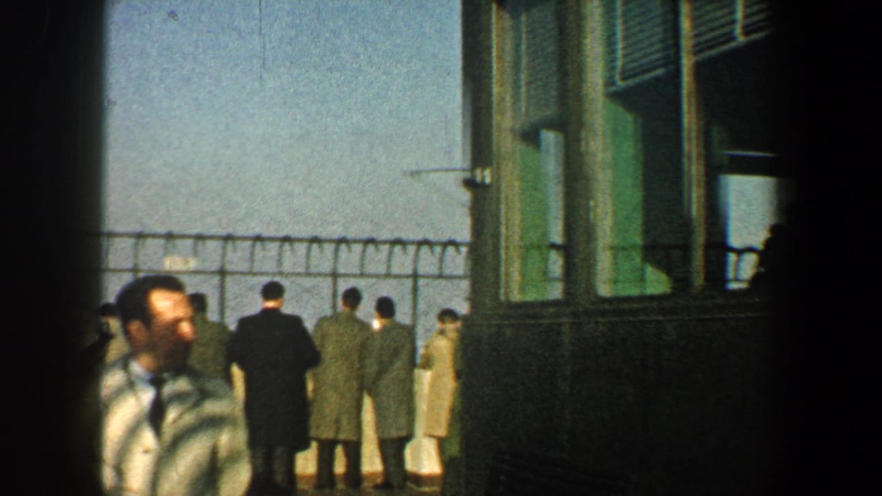 Tourists stand along a fenced observation deck while a striped-shirted man passes in the foreground.