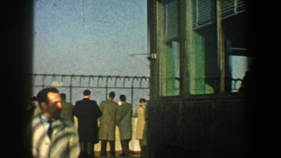 1960 New York City home-movie frame showing visitors at a fenced observation deck with a striped-shirted man near the camera.