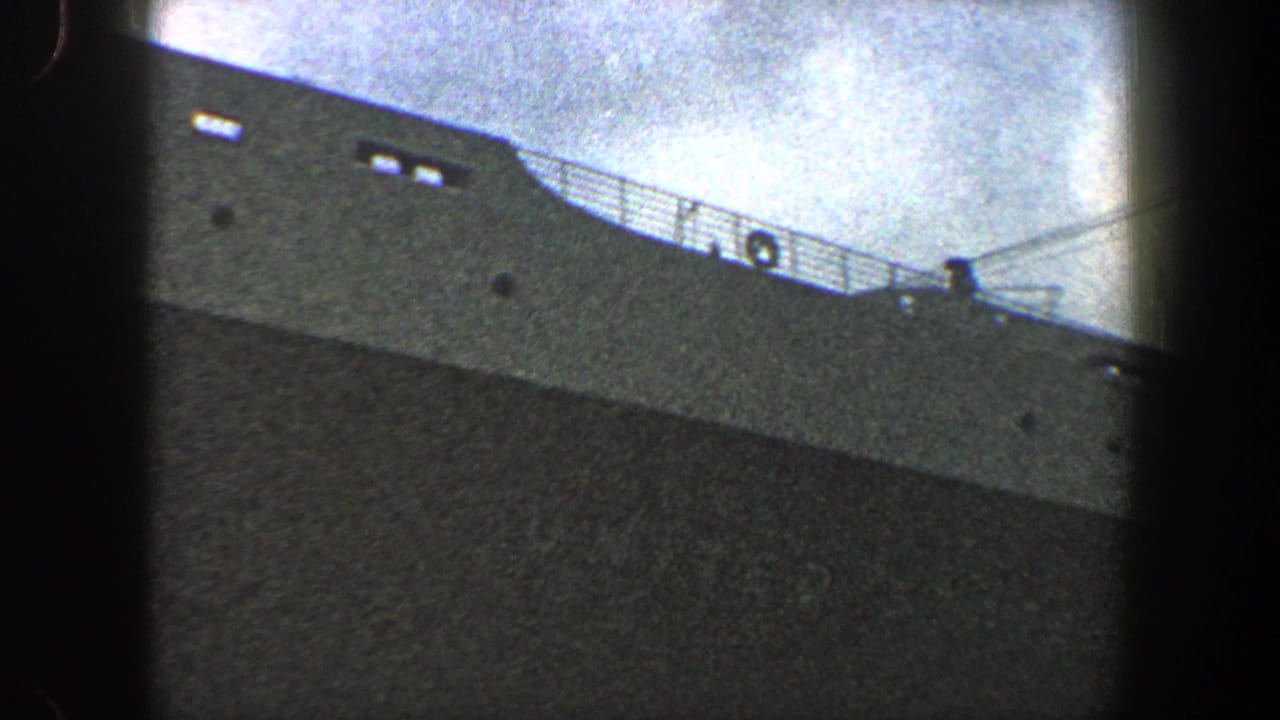 Upper portion of a large gray ship with railings and small dark openings.