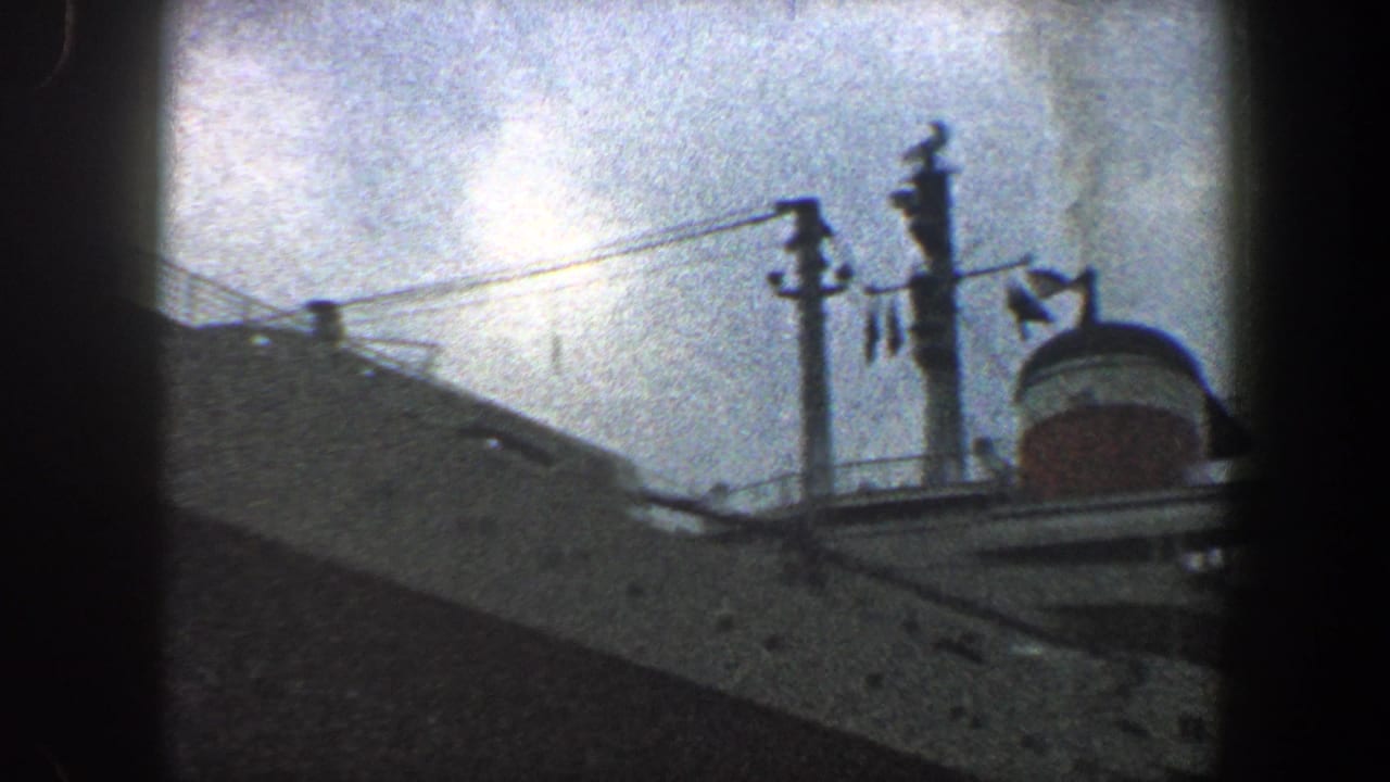 1960 New York City home-movie frame showing an ocean liner's upper hull, red-and-white funnel, railings, and masts from below.