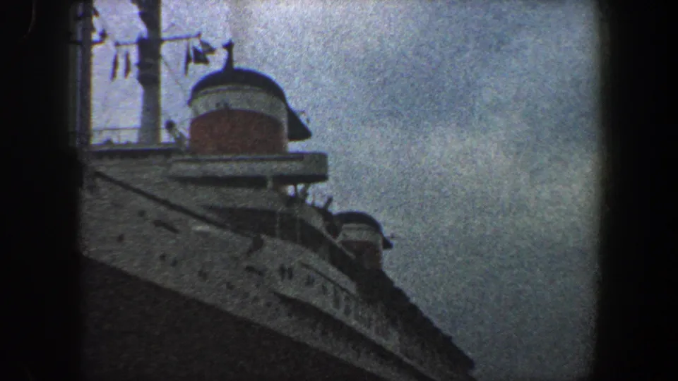 1960 New York City frame showing a passenger liner's curved hull with portholes, a red-and-white funnel, and deck railings under cloudy light.