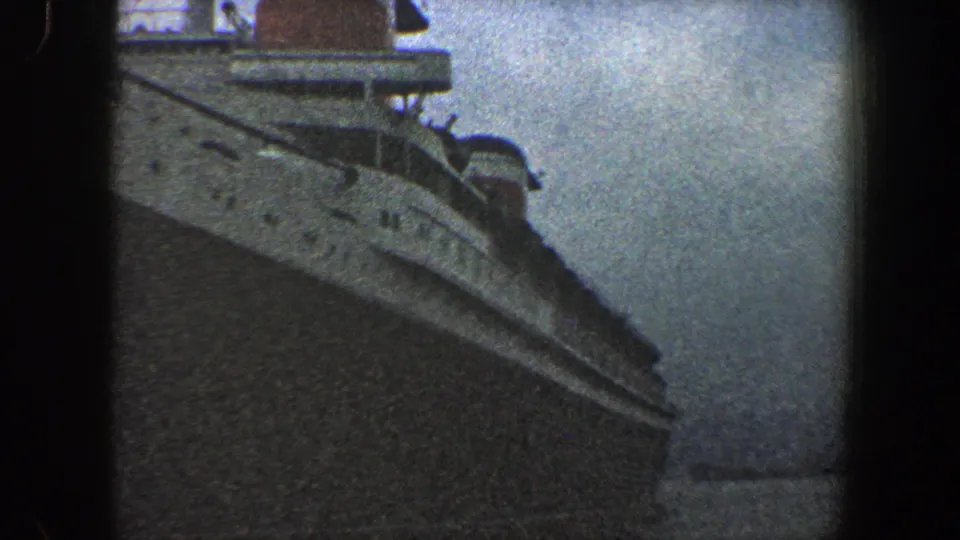 1960 New York City home-movie frame looking along the ocean liner's side, with portholes, railings, funnel, and overcast sky visible.