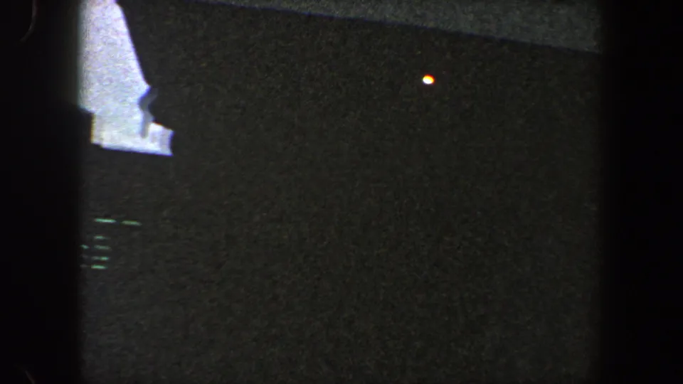Opening 8mm frame from 1960 New York City footage, mostly dark foreground with a small orange point of light and pale haze beyond.