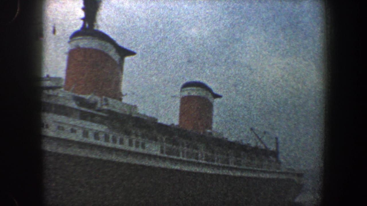 1960 New York City harbor home-movie frame showing an ocean liner's upper decks and red funnels beneath a gray sky.