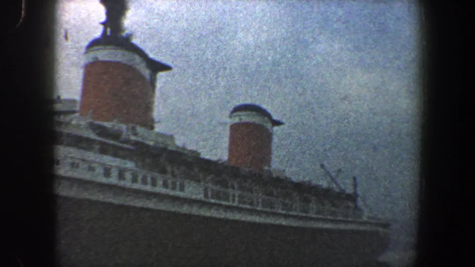 A grainy 8mm frame shows the ship's long upper deckline and two red funnels as the camera pans along the vessel.