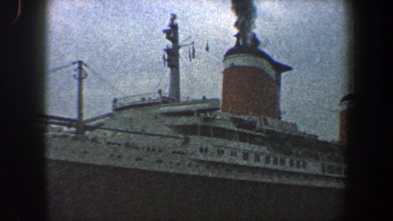 1960 New York City home-movie frame showing a large docked ocean liner with white upper decks, dark hull, and a smoking red-and-white funnel.