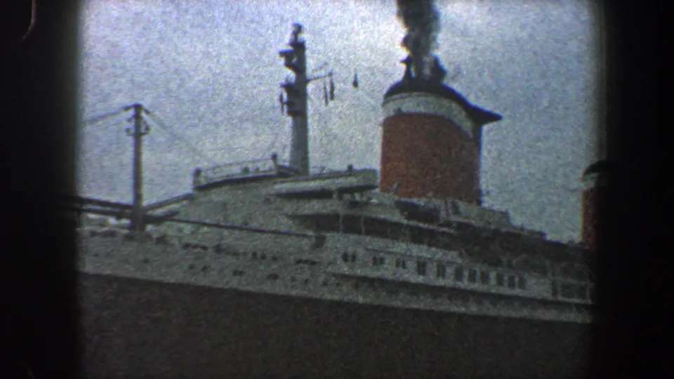 1960 New York City home-movie frame showing a large docked ocean liner with white upper decks, dark hull, and a smoking red-and-white funnel.