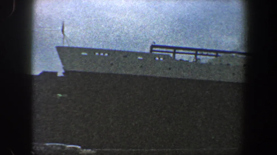 The 1960 New York City footage reaches the liner's forward section, with the bow area, deck railings, portholes, and dark hull visible.