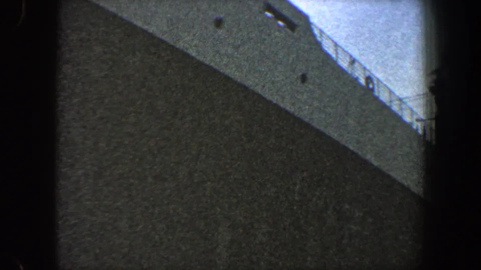 1960 8mm frame of a large vessel from below, with the dark hull dominating and a section of deck railing visible at right.