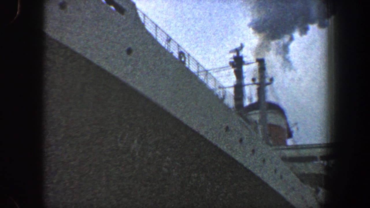 1960 New York City home-movie frame showing the bow and upper railings of an ocean liner, with dark smoke rising into a cloudy sky.