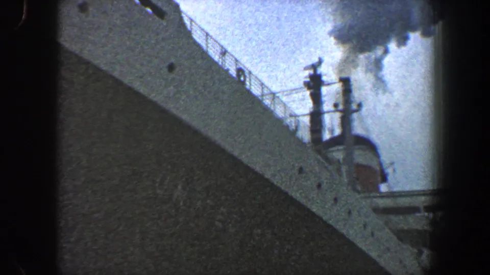 1960 New York City home-movie frame showing the bow and upper railings of an ocean liner, with dark smoke rising into a cloudy sky.